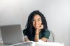 Joyful businesswoman with curly hair smiling at camera while using laptop indoors.